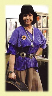 Woman in a purple blouse and beige pants posing indoors.